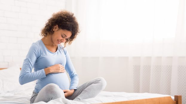 Mom expecting baby. Cute afro pregnant woman caressing her belly, sitting on bed, free space